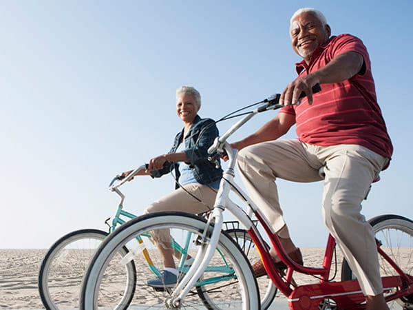 A senior Black couple is riding bicycles by a beach.