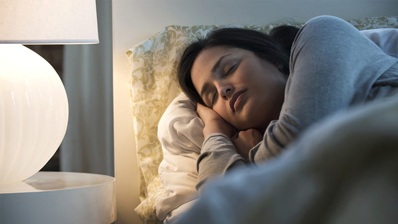 An Hispanic/Latina woman illuminated by a lamp is sleeping in bed.