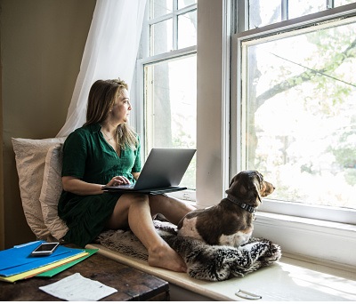 A young woman on a laptop looking out a bedroom window with her dog.