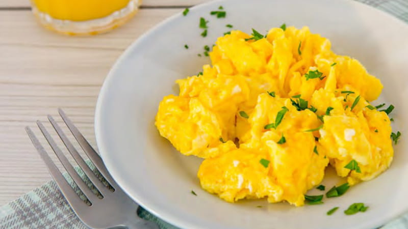A bowl of scrambled eggs sprinkled with parsley and a fork are on a table.