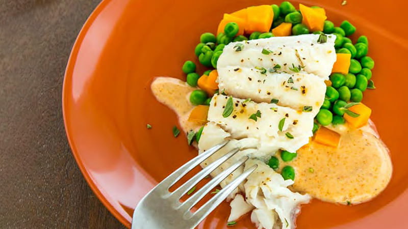 an orange plate of steamed catfish on a bed of peas, carrots and sauce with a fork digging in