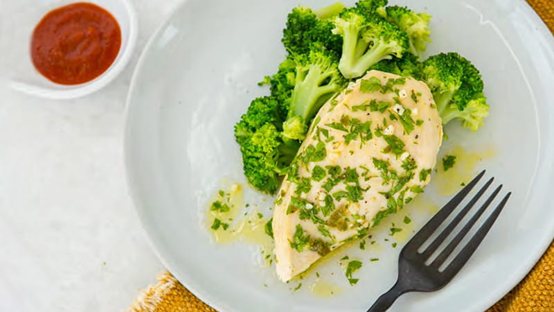 an aerial view of a plate of a steamed chicken breast sprinkled with parsley flakes, broccoli, and a side of sauce