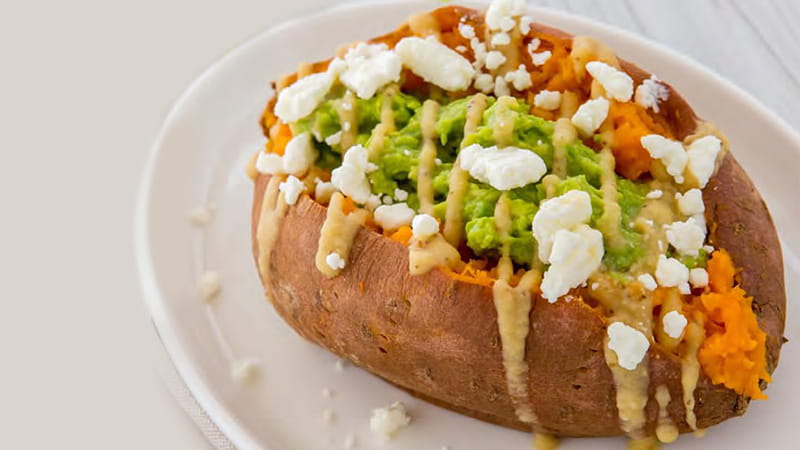 A whole sweet potato stuffed with avocado, feta cheese and drizzled with hummus dressing is on a plate.