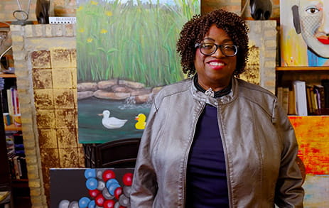 Stroke survivor, Minnie, is standing in a studio surrounded by her paintings.