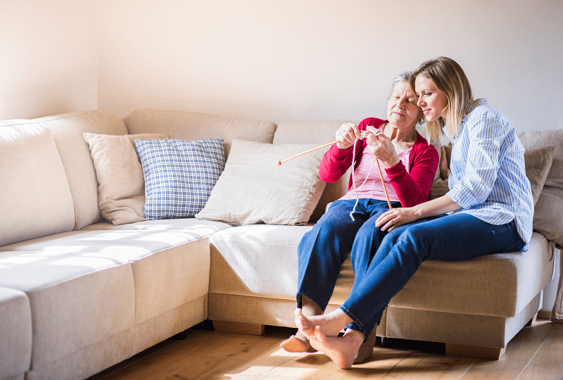 An older adult woman is sitting on a sofa knitting with a young adult woman.