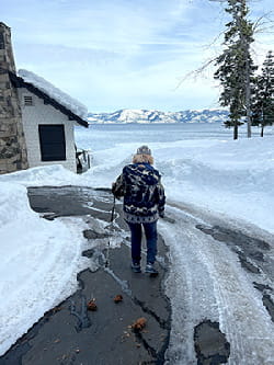 Deb Meyerson lleva abrigo y sombrero, y camina con bastón por un camino nevado con el lago Tahoe y las montañas de fondo